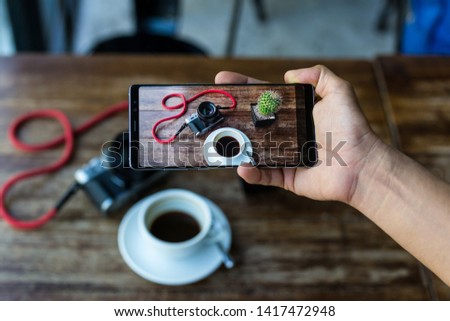 Blogger hand holding smart phone taking photo of coffee and camera on table.