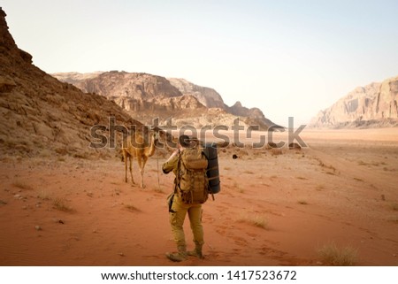 Male tourist in military brown clothing and large backpack taking picture of camel in wadi rum desert. Rocks, sand, dry grass and sandstone mountain ridge on background.