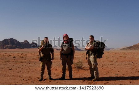 Three male friends posing together in Wadi Rum desert. Heavy big backpacks, military clothing, red keffiyeh, army boots on them. Sand, rocks, dry grass and sandstone mountain range on background.