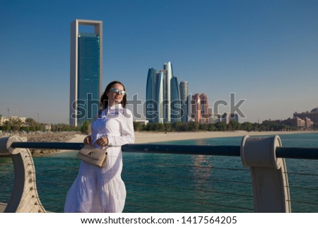 Beautiful brunette woman, has white blouse and long skirt, beige leather handbag, enjoying the views quay Abu Dhabi city in United Arab Emirates. Summer fashion style.