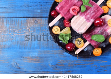 Fruit and berry popsicles on the plate with ice, pieces of kumquat, lime and cherries and mint against the blue background. Copy space