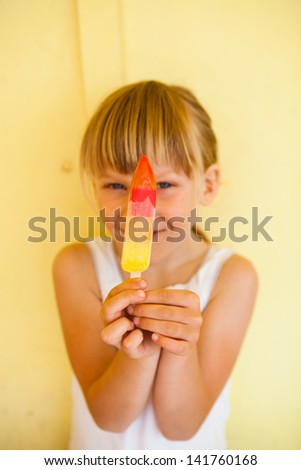 Smiling young girl holding up multicolored popsicle against yellow colored background