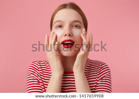 Cute young red-haired woman in a red striped T-shirt, with red lips , touches his face with fingers, looks at the camera and expects magical action of patches from dark circles under his eyes.