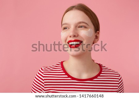 Close up of attractive young smiling red-haired woman with red lips and with patches under the eyes, wears in a red striped T-shirt, looks away with a happy expression, stands over pink background.