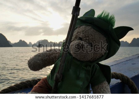 Beautiful Landscape Of Halong Bay and Teddy Bear In Green Dragon Costume On The Boat, Vietnam, Southeast Asia. UNESCO World Heritage Site. Grey-Blue Nature And White Light Of Sunset. Dark Background.