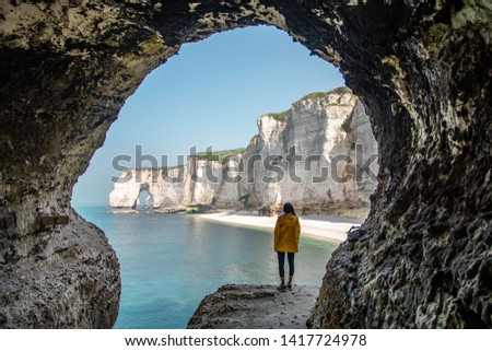 Landscape photo of a woman looking in the distance standing at the entrance of a circular cave with big cliffs, the beach and the ocean in the background. Shot in Etretat, Normandy, France. 
