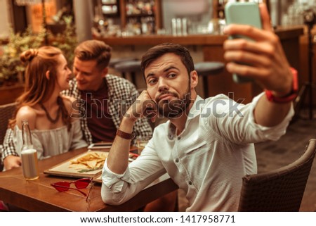 Taking selfies. Upset bored sad pessimistic sorrowful blue dark-haired nice-looking bearded man in his 25s taking selfies while his friends hugging at the background