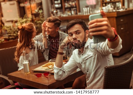 Taking photos. Upset pessimistic bored sad short-haired good-looking bearded male in his 25s taking selfies while his mates cuddling at the background
