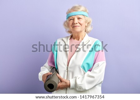 Happy elegant mature lady looking at camera while keeping yoga mat in hands, posing isolated over purple background.