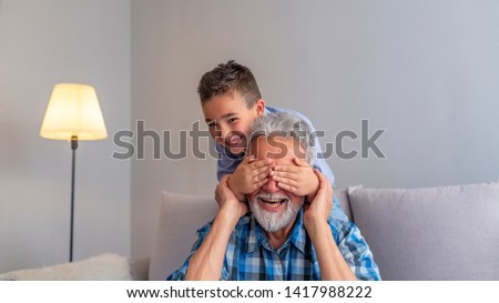 Portrait of handsome grandpa and grandson on gray background. Boy is covering grandpa's eye making a surprise. Grandfather and grandson playing and smiling at home 