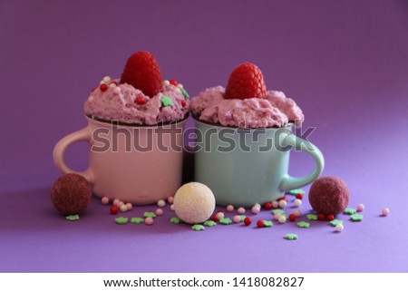 A pink and a blue cup of coffee with pink cream and raspberries on top, with pralines and sprinkle arround on lilac background