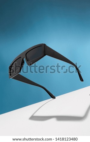 Front view shot of rectangular-shaped sunglasses with black rim and lenses. The tilted accessory is isolated in the air against the background with wide white stripe. Fashionable summer fashion item.