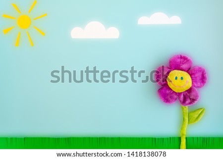Paper sun, clouds, green grass and toy big flower with a smiling face on a blue background. Funny summer landscape composition with a luminous effect on the theme of good sunny weather and good mood.