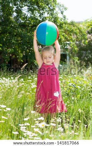 Little blond girl in a red summer dress is playing with a beach ball on a meadow full of blooming daisies