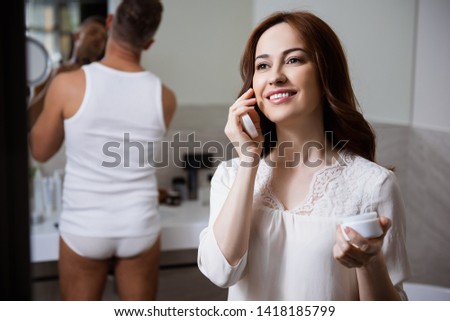 Selective focus of a young cheerful lady looking away with a smile while standing with a jar of cream in her hand. Man in underwear and a sleeveless shirt on the background