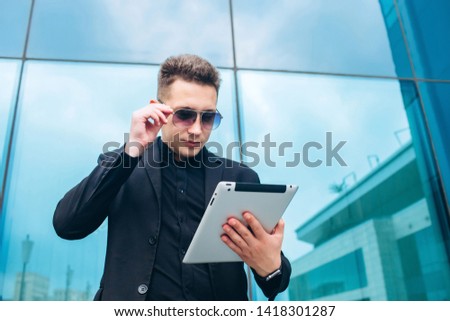 The guy in the black suit on the background of the glass modern business center. confident Businessman talking on cell phone and holding a tablet in his hand against the building with a glass facade