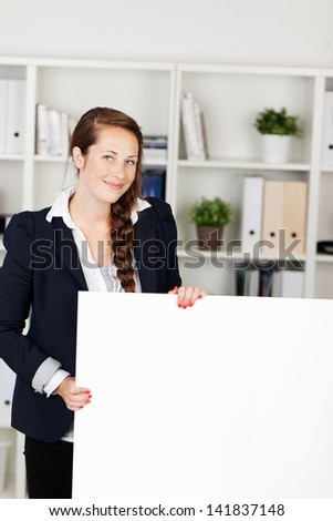 Beautiful young businesswoman with a friendly smile standing in an office holding a blank white placard for your text or advertisement