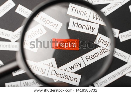 Magnifier glass over the red inscription inspire cut out of paper. Surrounded by other inscriptions on a dark background. Word cloud concept.