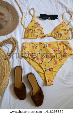 Feminine yellow floral bathing suit, sunglasses, brown mules, straw bag and hat. Flat lay, white linen background.