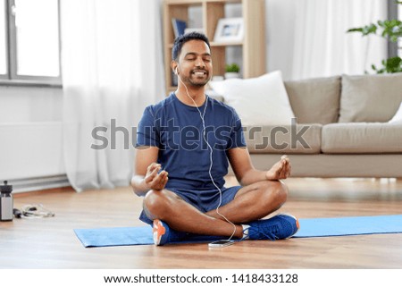 fitness, meditation and healthy lifestyle concept - indian man in earphones listening to music on smartphone and meditating in lotus pose at home Royalty-Free Stock Photo #1418433128