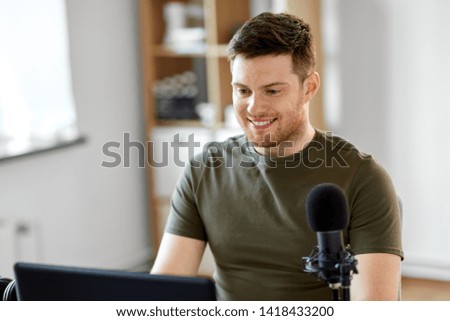 technology, mass media and podcast concept - happy young male audio blogger with laptop computer and microphone broadcasting at home office