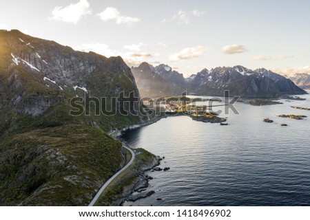 drone shot of a scenic village in front of massive mountains with sharp and narrow peaks in a beautiful landscape with calm fjord water of the ocean in front with beautiful sunlight taken from above