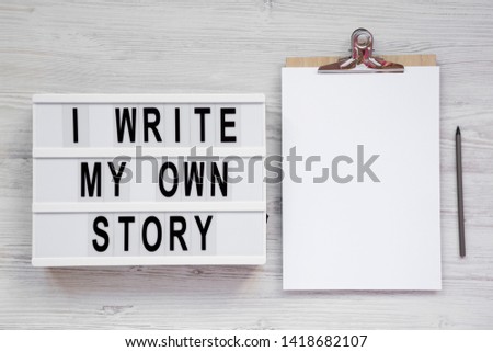 'I write my own story' words on a lightbox, clipboard with blank sheet of paper on a white wooden surface, top view. From above, overhead, flat lay.