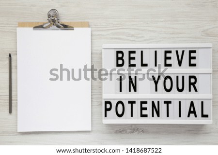 'Believe in your potential' words on a lightbox, clipboard with blank sheet of paper on a white wooden surface, top view. Close-up.
