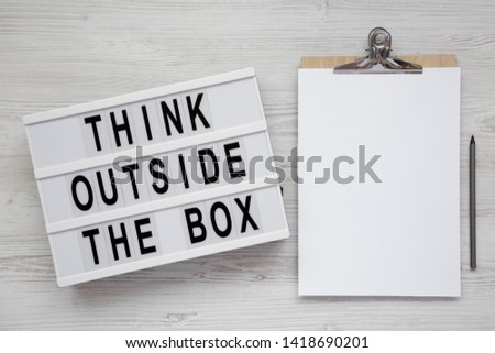 'Think outside the box' words on a light box, clipboard with blank sheet of paper on a white wooden background, top view. Flat lay, overhead, from above. Copy space.