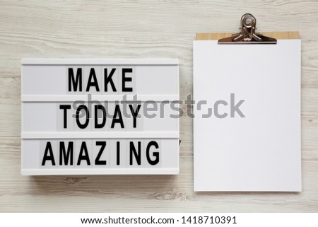 'Make today amazing' words on a light box, clipboard with blank sheet of paper on a white wooden background. From above, overhead, flat lay, top view.