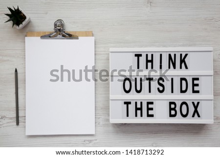 'Think outside the box' words on a light box, clipboard with blank sheet of paper over white wooden surface, top view. Flat lay, overhead, from above. 