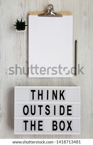 'Think outside the box' words on a light box, clipboard with blank sheet of paper over white wooden background, top view. Flat lay, overhead, from above. 