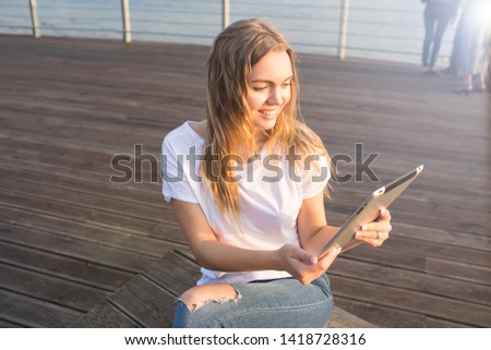 Happy smiling woman online ordering via portable touch pad while relaxing on embankment during recreation time in weekend. Joyful female watching funny video in social network via digital gadget 