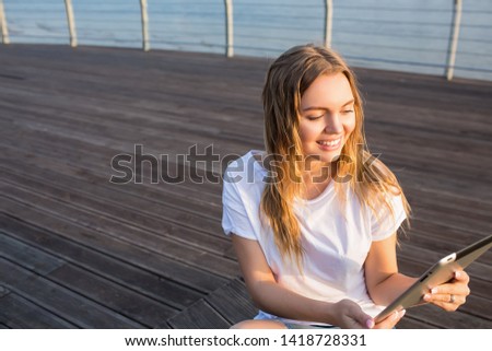 Happy smiling woman skilled lifestyle blogger online ordering via touch pad computer while relaxing outdoors on wooden embankment near copy space for promotional content. Joyful female using gadget 