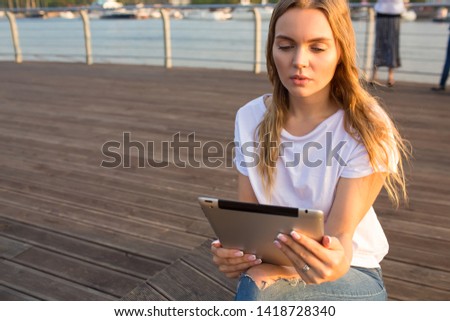 Gorgeous woman online booking via portable touch pad gadget while relaxing outdoors during summer vacations. Thoughtful college female reading e-book via digital tablet, resting on quay in sunny day