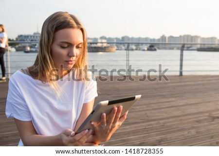 Young blonde caucasian woman smart university student reading article via touch pad computer while relaxing on wooden embankment near copy space for your promotional content. Female online ordering 