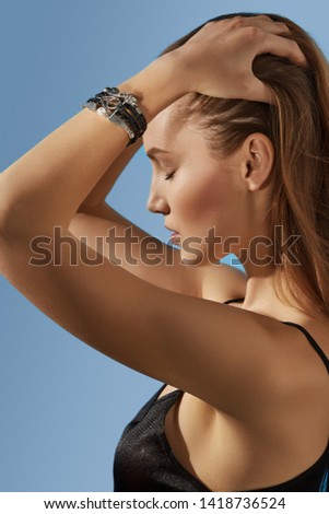 Cropped side shot of lady, wearing crop top and leather multi-strand bracelet with various silver inserts in view of wings and owls. The woman is fixing hair with eyes closed over blue background.