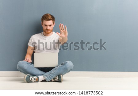 Young redhead student man sitting on the floor putting hand in front. He is holding a laptop.