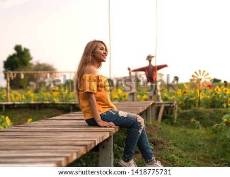 Asian tanned woman wearing a pretty yellow top sitting on a wooden walkway flower plantation - Young Thai female smiling and happy with bright sunshine in a sunflower farmers field with scarecrows