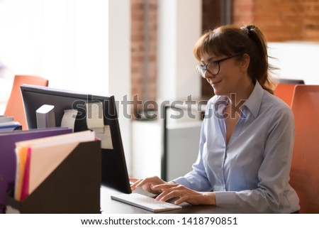 Middle aged employee businesswoman working typing on keyboard business e-mail looking at pc screen feels satisfied alone at modern shared coworking space. Solve matters distantly busy workday concept