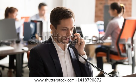 Serious company employee male in formal suit sitting in shared office room coworking space with other workers talking on landline phone with client. Call centre support distance communication concept Royalty-Free Stock Photo #1418790872