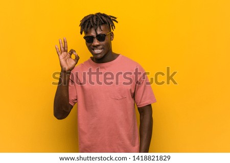 Young casual black man wearing sunglasses winks an eye and holds an okay gesture with hand.