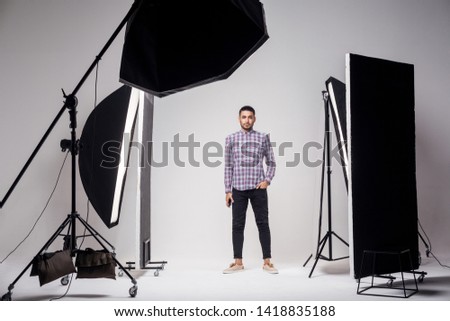 Professional photography studio showing behind the scenes lights. fashion handsome young man model at studio in the light flashes, standing and looking at camera. indoor studio shot on grey background Royalty-Free Stock Photo #1418835188
