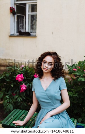 caucasian fashion model with curly hairstyle and make up posing on a bench near the house with fair walls, blue dress, summer, soft light, peonies