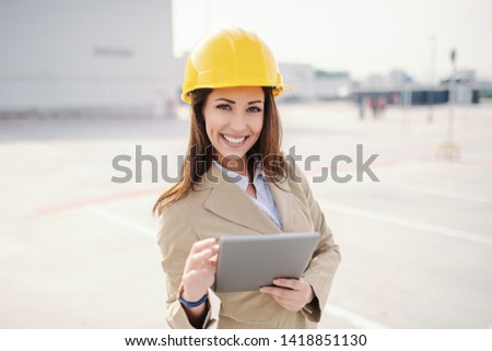 Attractive Caucasian female architect dressed smart casual and with helmet on head using tablet while standing on construction site.