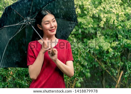 Young asian woman wearing red T-shirt smiling under black umbrella in the rain, Green nature background, Rainy season.