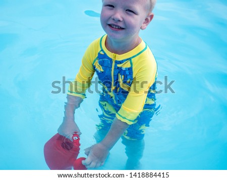 Little blonde boy  in hot summer day swim in carcas pool  in swimwear for boys. Kid is holding in hand red watering can