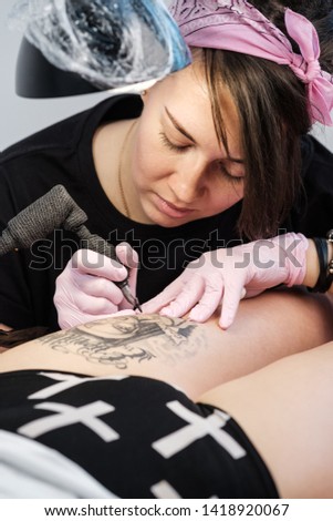 Female tattoo master tattooing female client. Figure nuns on the thigh. Black tattoo machine and lamp. Workplace artist. White background.