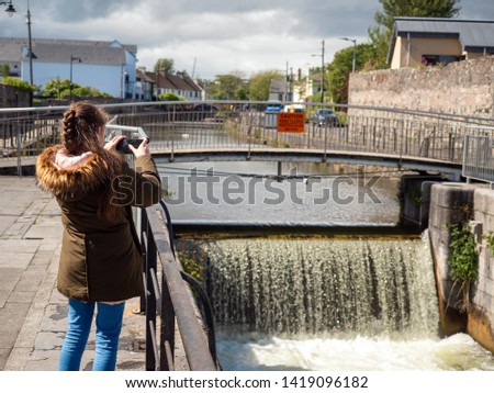 Young girl taking picture on her smart phone of a water falling in a dam. Sunny day, Town environment.