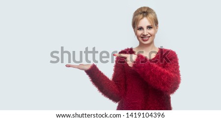 Portrait of happy beautiful young blond woman in red blouse standing, looking at camera with toothy smile, pointing at something holding on hand. indoor studio shot isolated on light gray background.